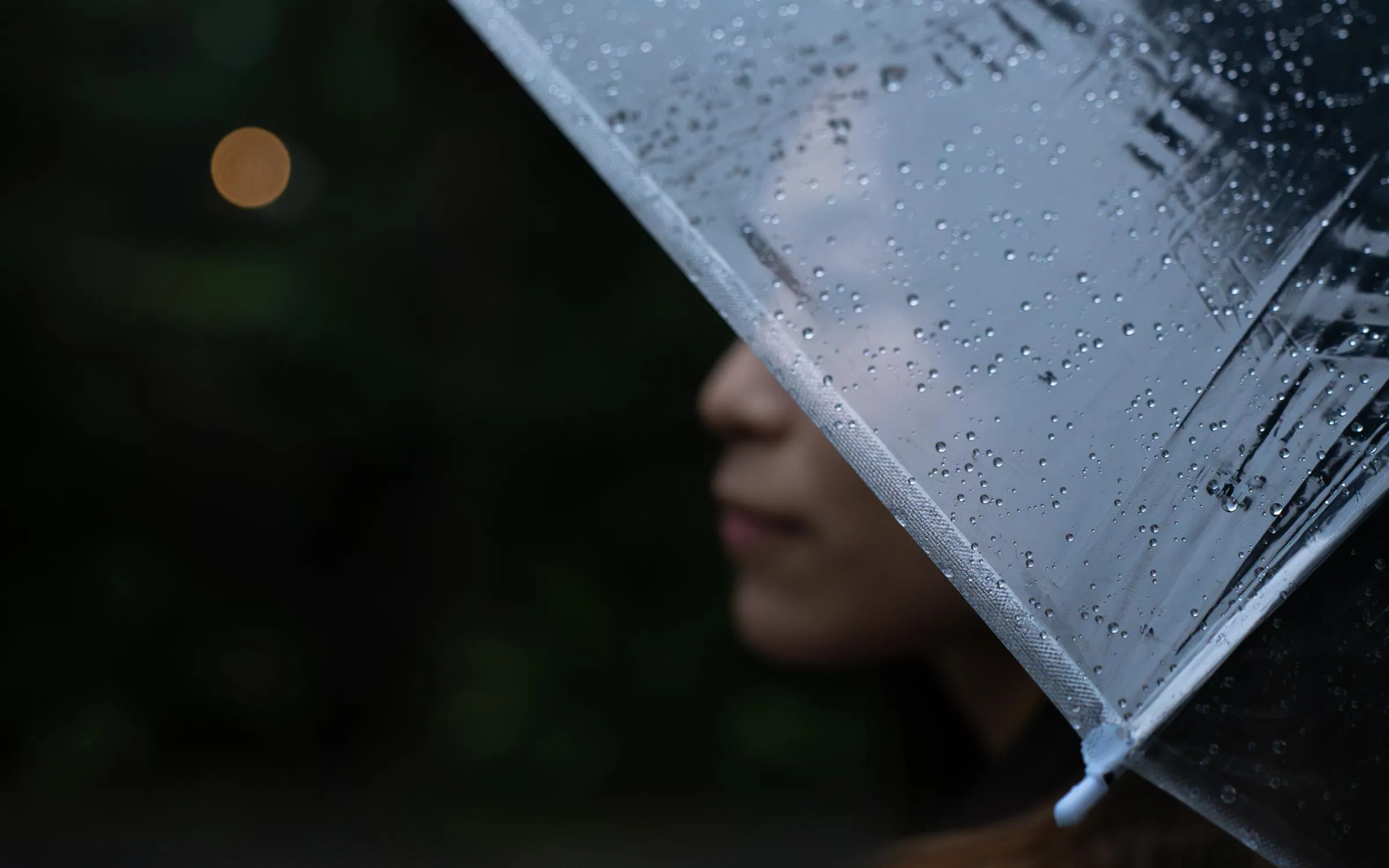 Under a clear umbrella in the rain, a quiet gaze into the distance, streetlights melting into soft golden bokeh