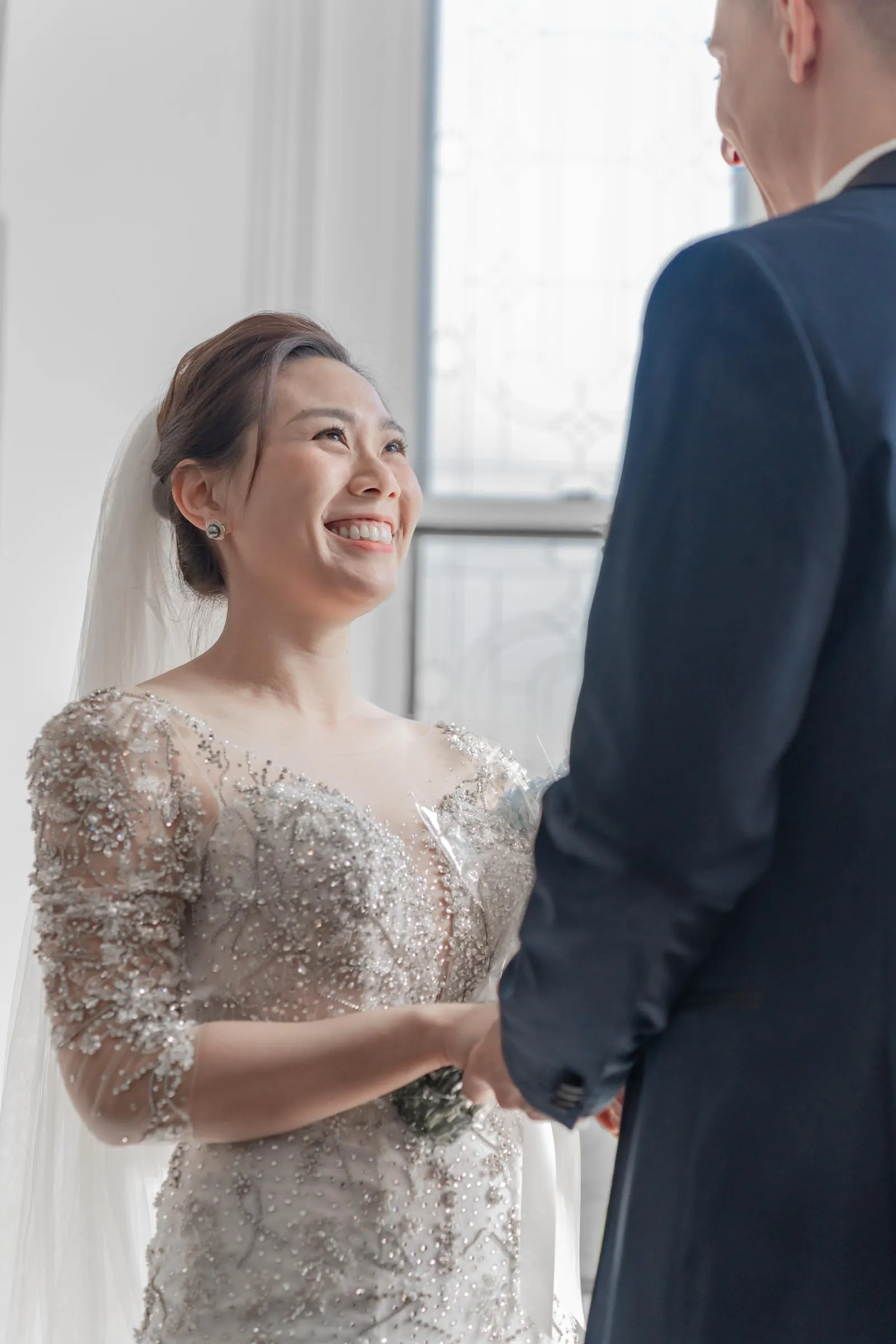 The bride looks up at her beloved with a smile she cannot hold back, bathed in the soft glow by the window.